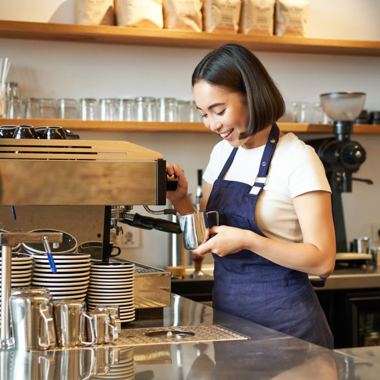 Happy asian girl barista, working in cafe, brewing coffee, prepare cappuccino, using steamer on machine to prepare latte, smiling and laughing.