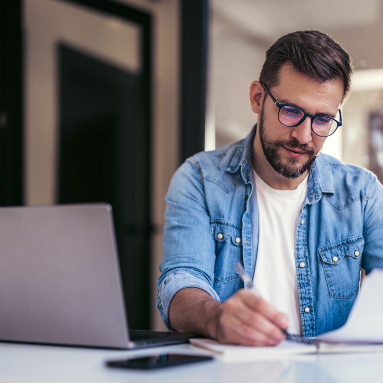 Man reading paper documents, working with laptop at workplace, sitting at desk.