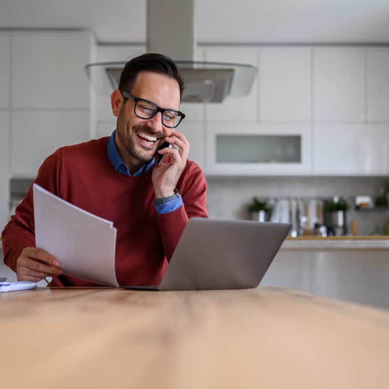 Happy young businessman holding documents and talking over smart phone while working from home
