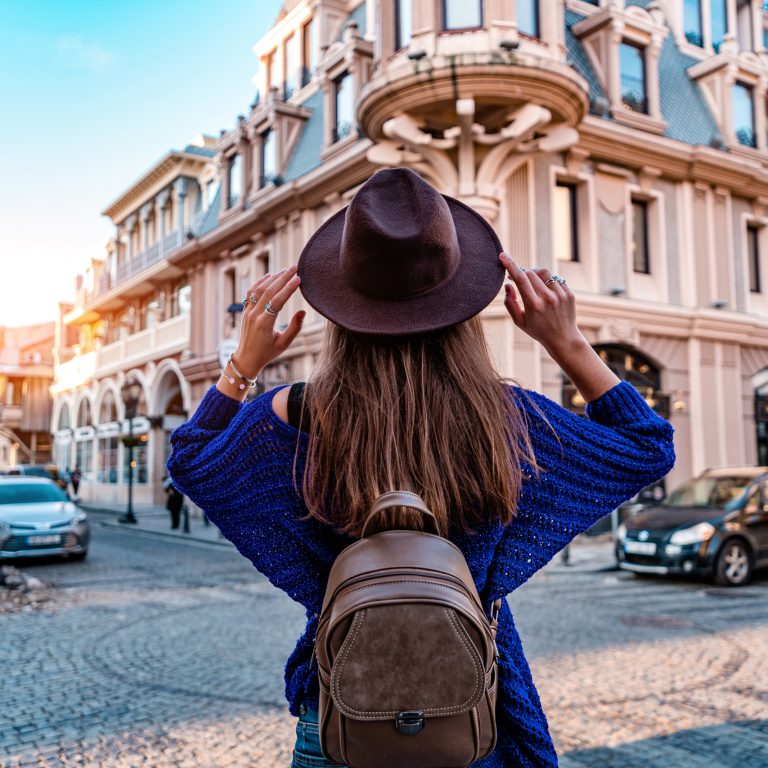 Casual stylish fashionable hipster traveler woman in hat and with brown backpack walking alone around european city