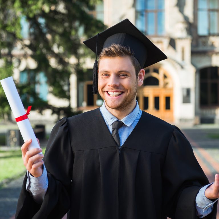Young male student dressed in black graduation gown. Campus as a background. Boy cheerfully smiling, holding diploma and looking at camera